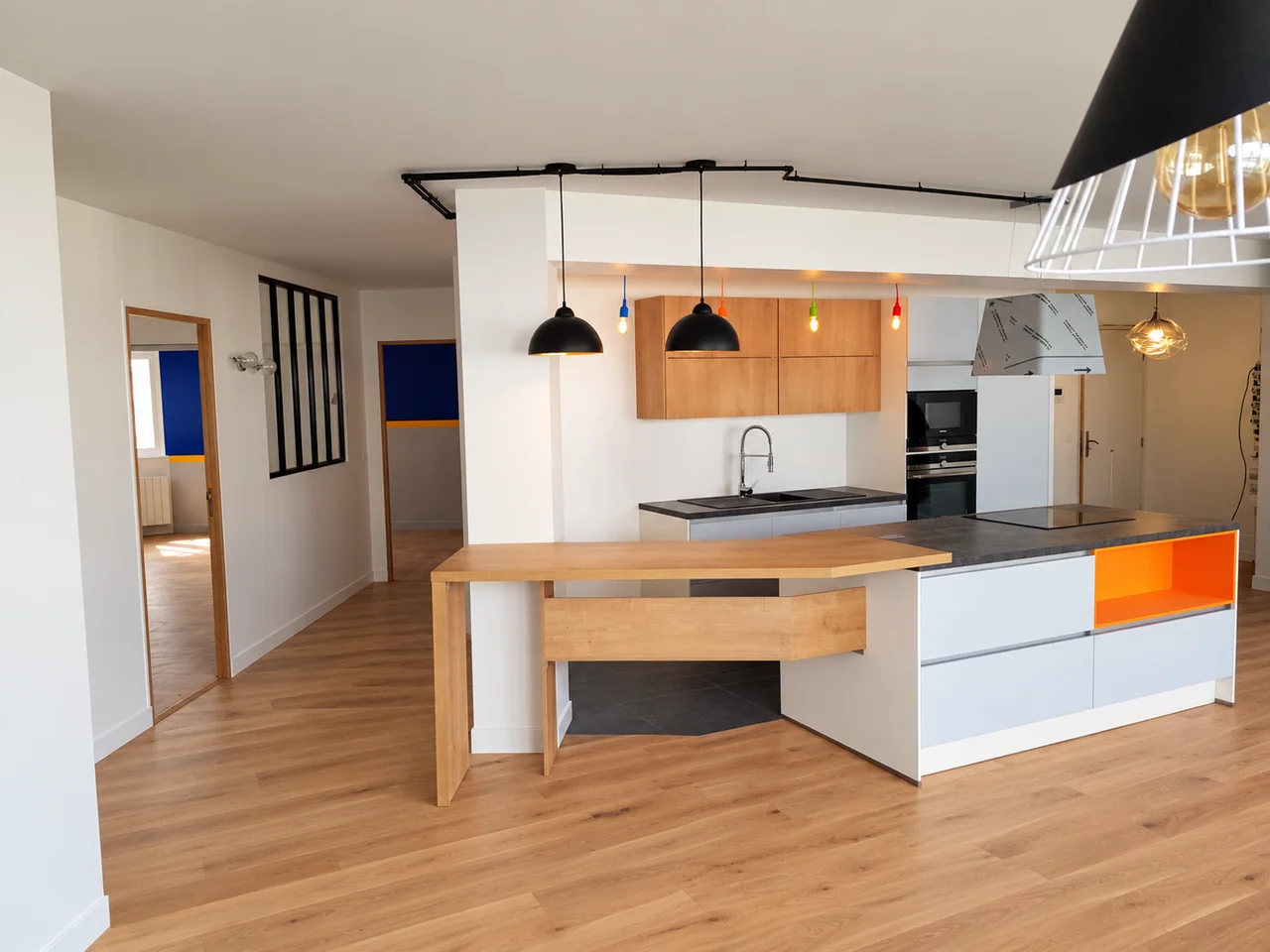 Cuisine avec îlot blanc, niche orange, tablette aspect bois et suspensions noires.