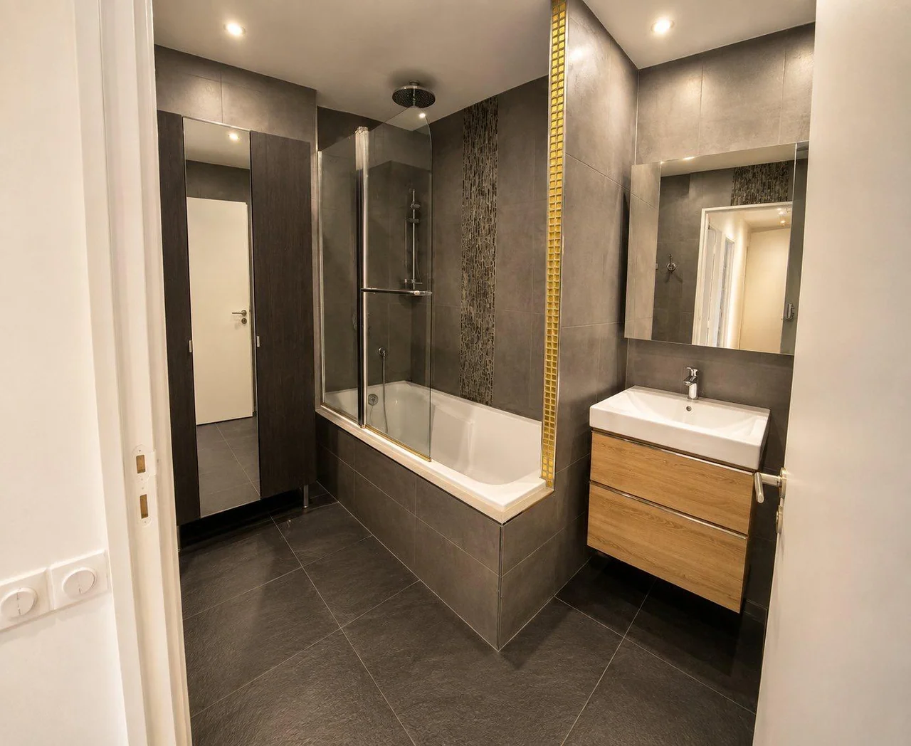 Salle de bains carrelée en gris avec baignoire, paroi vitrée et meuble vasque aspect bois.