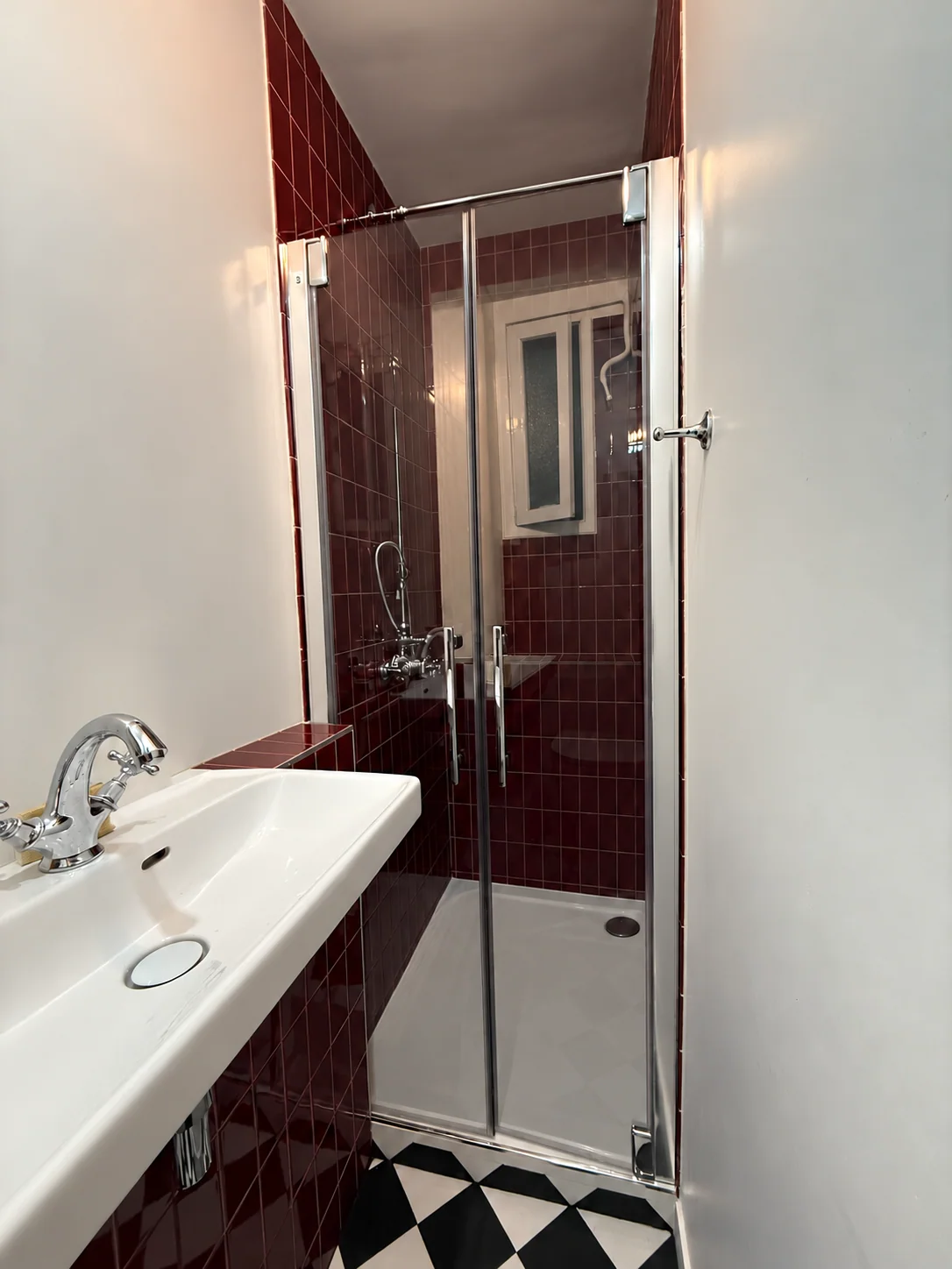 Douche vitrée et lavabo blanc avec carrelage bordeaux et sol noir et blanc.