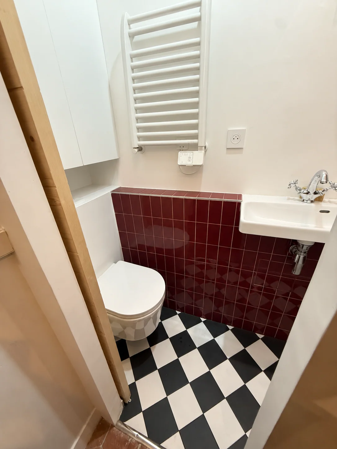 WC blanc et lavabo dans une salle d’eau au carrelage bordeaux et au sol en damier noir et blanc.
