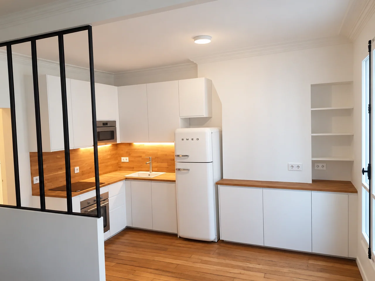 Cuisine blanche en L avec plan aspect bois, réfrigérateur blanc et verrière noire.